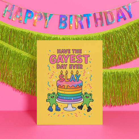 5x7 inch birthday greeting card with a yellow background featuring the phrase “Have the Gayest Day Ever” in bold pink text, a rainbow birthday cake with candles spelling “SLAY,” and two green frogs wearing colorful platform boots. Card includes confetti details and a white envelope. Printed on 120lb silk cover stock. The inside message reads: “Sashay into this new age with zero regrets and maximum glitter.”