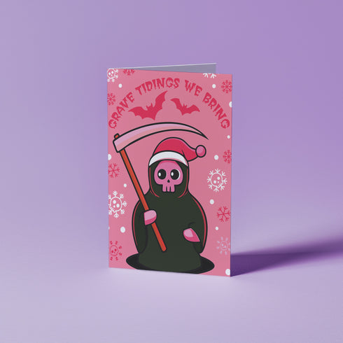 Quirky Christmas card featuring grim reaper in Santa hat. Pink background with bats, snowflakes, skull motifs. Text reads 'Grave Tidings We Bring'. Humorous blend of holiday cheer and gothic aesthetic. Perfect for alternative Christmas greetings.