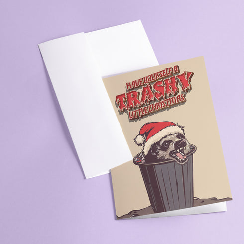 A playful holiday card featuring a raccoon wearing a Santa hat, popping out of a trash can with a mischievous grin. The text above reads "Have Yourself a Trashy Little Christmas" in bold, dripping letters. The card combines humor with a grungy, festive theme for a lighthearted take on the holiday season.