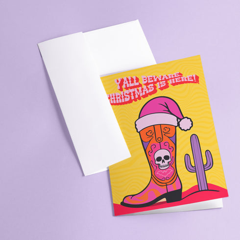 Colorful Christmas card with a yellow background. Features a stylized cowboy boot decorated with a skull, hearts, and swirls in pink, orange, and purple. The boot wears a pink Santa hat. A green cactus stands beside the boot. Top text reads 'Y'ALL BEWARE, CHRISTMAS IS HERE!' in red, wavy letters. Card has a purple border. Inside message: 'May Your days be merry, bright and full of southern delight.' The design blends Christmas, Western, and Day of the Dead motifs for a unique, playful holiday greeting