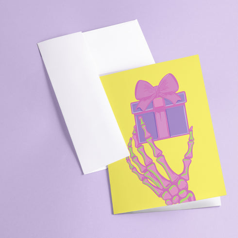 Birthday card with vibrant yellow background. A pink and green skeletal hand holds a purple and pink gift box with large pink bow. The illustration blends festive and spooky elements, creating a playful Halloween-meets-birthday aesthetic. The stylized, colorful design uses contrasting bright colors for a eye-catching effect. Inside message reads: 'Unwrap the Magic. Embrace the Spook. It's Your Day to Shine!' - combining celebratory spirit with a touch of the macabre for a unique birthday greeting.