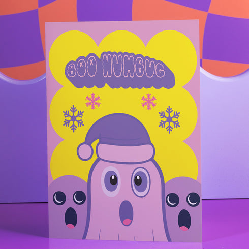 Christmas card with a playful ghost theme. Central cute pink ghost wearing a purple Santa hat, flanked by two smaller purple ghosts. Yellow background with snowflakes. Text reads "Boo HUMBUG" in purple bubble letters. Blends Christmas and Halloween elements for a unique holiday greeting. Inside message: "May your Christmas be filled with delight, not fright.