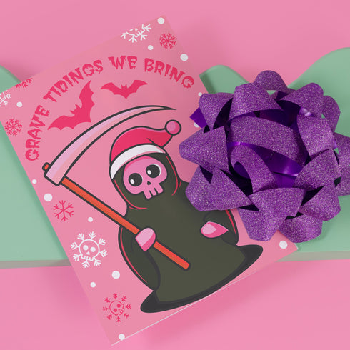Quirky Christmas card featuring grim reaper in Santa hat. Pink background with bats, snowflakes, skull motifs. Text reads 'Grave Tidings We Bring'. Humorous blend of holiday cheer and gothic aesthetic. Perfect for alternative Christmas greetings.