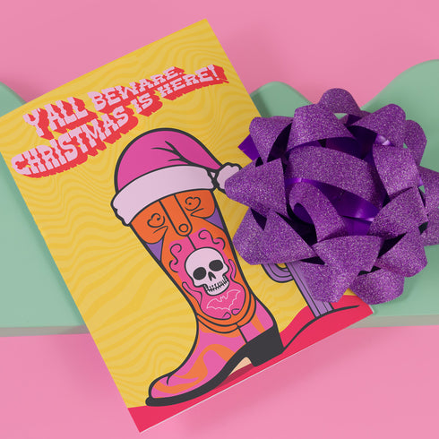 Colorful Christmas card with a yellow background. Features a stylized cowboy boot decorated with a skull, hearts, and swirls in pink, orange, and purple. The boot wears a pink Santa hat. A green cactus stands beside the boot. Top text reads 'Y'ALL BEWARE, CHRISTMAS IS HERE!' in red, wavy letters. Card has a purple border. Inside message: 'May Your days be merry, bright and full of southern delight.' The design blends Christmas, Western, and Day of the Dead motifs for a unique, playful holiday greeting
