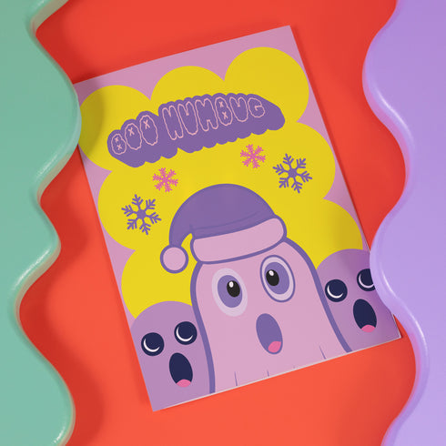 Christmas card with a playful ghost theme. Central cute pink ghost wearing a purple Santa hat, flanked by two smaller purple ghosts. Yellow background with snowflakes. Text reads "Boo HUMBUG" in purple bubble letters. Blends Christmas and Halloween elements for a unique holiday greeting. Inside message: "May your Christmas be filled with delight, not fright.