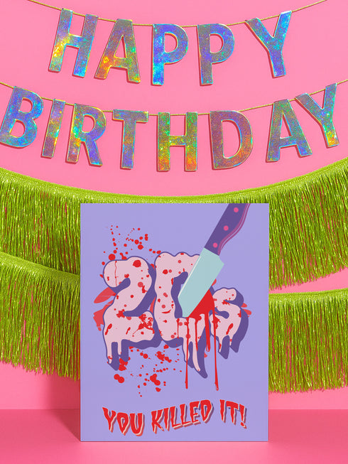 A 5x7 birthday card with a horror-comedy design. The front features the word "20s" in pink gooey lettering with a bloody knife stabbing through it, surrounded by red blood splatter. Below it reads "YOU KILLED IT!" in dripping red font, set against a purple background with a rainbow foil tinsel curtain behind it. Inside, the right panel says "HERE'S TO THE NEXT VICTIM: YOUR THIRTIES!" with dripping red blood at the top and a pink cherry garland background.
