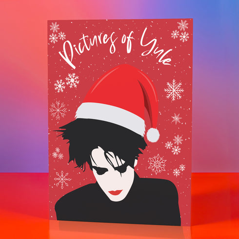 A festive greeting card featuring an illustration of Robert Smith from The Cure, wearing a Santa hat. The card has a red background with white snowflakes and the text "Pictures of Yule" at the top, playfully referencing the song "Pictures of You." The card merges gothic and holiday themes for a unique seasonal twist.