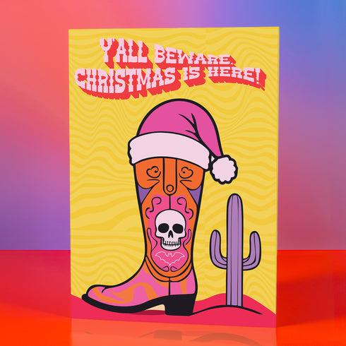 Colorful Christmas card with a yellow background. Features a stylized cowboy boot decorated with a skull, hearts, and swirls in pink, orange, and purple. The boot wears a pink Santa hat. A green cactus stands beside the boot. Top text reads 'Y'ALL BEWARE, CHRISTMAS IS HERE!' in red, wavy letters. Card has a purple border. Inside message: 'May Your days be merry, bright and full of southern delight.' The design blends Christmas, Western, and Day of the Dead motifs for a unique, playful holiday greeting