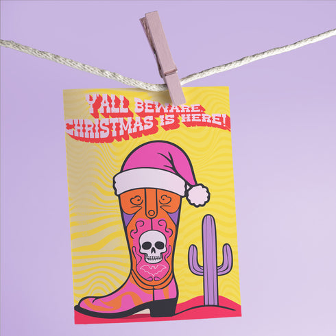 Colorful Christmas card with a yellow background. Features a stylized cowboy boot decorated with a skull, hearts, and swirls in pink, orange, and purple. The boot wears a pink Santa hat. A green cactus stands beside the boot. Top text reads 'Y'ALL BEWARE, CHRISTMAS IS HERE!' in red, wavy letters. Card has a purple border. Inside message: 'May Your days be merry, bright and full of southern delight.' The design blends Christmas, Western, and Day of the Dead motifs for a unique, playful holiday greeting