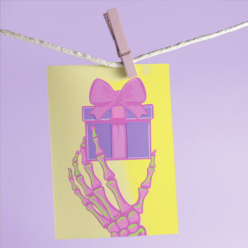 Birthday card with vibrant yellow background. A pink and green skeletal hand holds a purple and pink gift box with large pink bow. The illustration blends festive and spooky elements, creating a playful Halloween-meets-birthday aesthetic. The stylized, colorful design uses contrasting bright colors for a eye-catching effect. Inside message reads: 'Unwrap the Magic. Embrace the Spook. It's Your Day to Shine!' - combining celebratory spirit with a touch of the macabre for a unique birthday greeting.