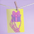 Birthday card with vibrant yellow background. A pink and green skeletal hand holds a purple and pink gift box with large pink bow. The illustration blends festive and spooky elements, creating a playful Halloween-meets-birthday aesthetic. The stylized, colorful design uses contrasting bright colors for a eye-catching effect. Inside message reads: 'Unwrap the Magic. Embrace the Spook. It's Your Day to Shine!' - combining celebratory spirit with a touch of the macabre for a unique birthday greeting.