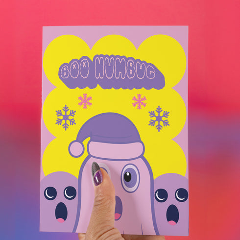 Christmas card with a playful ghost theme. Central cute pink ghost wearing a purple Santa hat, flanked by two smaller purple ghosts. Yellow background with snowflakes. Text reads "Boo HUMBUG" in purple bubble letters. Blends Christmas and Halloween elements for a unique holiday greeting. Inside message: "May your Christmas be filled with delight, not fright.