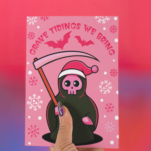 Quirky Christmas card featuring grim reaper in Santa hat. Pink background with bats, snowflakes, skull motifs. Text reads 'Grave Tidings We Bring'. Humorous blend of holiday cheer and gothic aesthetic. Perfect for alternative Christmas greetings.