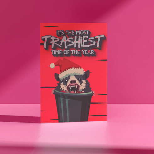 A humorous holiday card featuring a cartoon possum popping out of a trash can, wearing a Santa hat with fangs bared. The text at the top reads "It's the most trashiest time of the year," adding a playful and quirky twist to the holiday season.