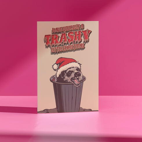 A playful holiday card featuring a raccoon wearing a Santa hat, popping out of a trash can with a mischievous grin. The text above reads "Have Yourself a Trashy Little Christmas" in bold, dripping letters. The card combines humor with a grungy, festive theme for a lighthearted take on the holiday season.