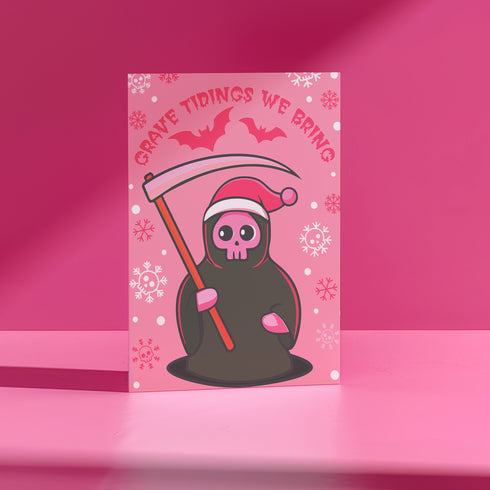 Quirky Christmas card featuring grim reaper in Santa hat. Pink background with bats, snowflakes, skull motifs. Text reads 'Grave Tidings We Bring'. Humorous blend of holiday cheer and gothic aesthetic. Perfect for alternative Christmas greetings.