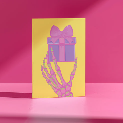 Birthday card with vibrant yellow background. A pink and green skeletal hand holds a purple and pink gift box with large pink bow. The illustration blends festive and spooky elements, creating a playful Halloween-meets-birthday aesthetic. The stylized, colorful design uses contrasting bright colors for a eye-catching effect. Inside message reads: 'Unwrap the Magic. Embrace the Spook. It's Your Day to Shine!' - combining celebratory spirit with a touch of the macabre for a unique birthday greeting.