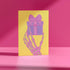 Birthday card with vibrant yellow background. A pink and green skeletal hand holds a purple and pink gift box with large pink bow. The illustration blends festive and spooky elements, creating a playful Halloween-meets-birthday aesthetic. The stylized, colorful design uses contrasting bright colors for a eye-catching effect. Inside message reads: 'Unwrap the Magic. Embrace the Spook. It's Your Day to Shine!' - combining celebratory spirit with a touch of the macabre for a unique birthday greeting.