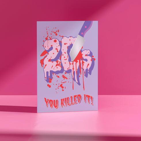 A 5x7 birthday card with a horror-comedy design. The front features the word "20s" in pink gooey lettering with a bloody knife stabbing through it, surrounded by red blood splatter. Below it reads "YOU KILLED IT!" in dripping red font, set against a purple background with a rainbow foil tinsel curtain behind it. Inside, the right panel says "HERE'S TO THE NEXT VICTIM: YOUR THIRTIES!" with dripping red blood at the top and a pink cherry garland background.