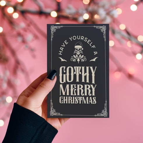 A black holiday card featuring gothic-style text that reads "Have Yourself a Gothy Merry Christmas." The design includes a skull wearing a Santa hat, bats, and ornate decorative borders, combining a dark, spooky aesthetic with festive holiday elements.