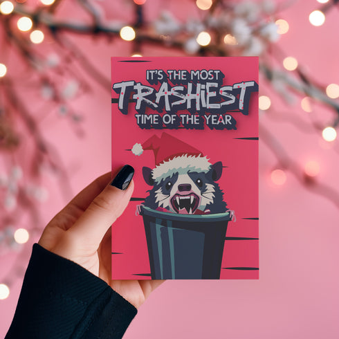 A humorous holiday card featuring a cartoon possum popping out of a trash can, wearing a Santa hat with fangs bared. The text at the top reads "It's the most trashiest time of the year," adding a playful and quirky twist to the holiday season.