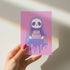 Birthday card featuring a cute cartoon grim reaper holding a birthday cake on pink background. The reaper wears a purple cloak and has a white skull face. It's carrying a small purple cake with three lit candles. The reaper's cloak drips like melting icing at the bottom. Ideal for Halloween birthdays or gothic-themed celebrations. Inside greeting: "Hope your B-day is spooky AF and sweet as cake!"