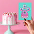 Quirky birthday card with teal background. Pink-purple cake topped by large cartoon eyeball. Three eyeball-shaped balloons float above, each with blue iris and pink edges. Inside message: "Eye Think You're Amazing. Happy Birthday". Playful design combines celebration with surreal humor, creating a memorable and slightly bizarre greeting. Whimsical imagery and pun offer a unique twist on traditional birthday wishes.