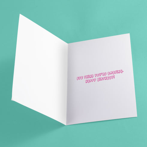 Quirky birthday card with teal background. Pink-purple cake topped by large cartoon eyeball. Three eyeball-shaped balloons float above, each with blue iris and pink edges. Inside message: "Eye Think You're Amazing. Happy Birthday". Playful design combines celebration with surreal humor, creating a memorable and slightly bizarre greeting. Whimsical imagery and pun offer a unique twist on traditional birthday wishes.