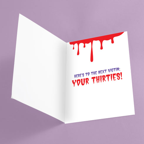 A 5x7 birthday card with a horror-comedy design. The front features the word "20s" in pink gooey lettering with a bloody knife stabbing through it, surrounded by red blood splatter. Below it reads "YOU KILLED IT!" in dripping red font, set against a purple background with a rainbow foil tinsel curtain behind it. Inside, the right panel says "HERE'S TO THE NEXT VICTIM: YOUR THIRTIES!" with dripping red blood at the top and a pink cherry garland background.
