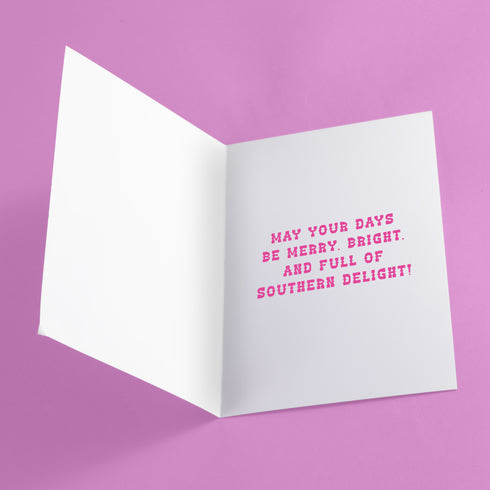 Colorful Christmas card with a yellow background. Features a stylized cowboy boot decorated with a skull, hearts, and swirls in pink, orange, and purple. The boot wears a pink Santa hat. A green cactus stands beside the boot. Top text reads 'Y'ALL BEWARE, CHRISTMAS IS HERE!' in red, wavy letters. Card has a purple border. Inside message: 'May Your days be merry, bright and full of southern delight.' The design blends Christmas, Western, and Day of the Dead motifs for a unique, playful holiday greeting