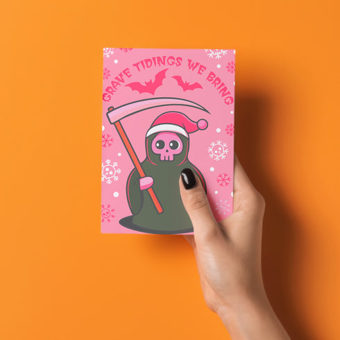 Quirky Christmas card featuring grim reaper in Santa hat. Pink background with bats, snowflakes, skull motifs. Text reads 'Grave Tidings We Bring'. Humorous blend of holiday cheer and gothic aesthetic. Perfect for alternative Christmas greetings.