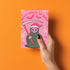 Quirky Christmas card featuring grim reaper in Santa hat. Pink background with bats, snowflakes, skull motifs. Text reads 'Grave Tidings We Bring'. Humorous blend of holiday cheer and gothic aesthetic. Perfect for alternative Christmas greetings.
