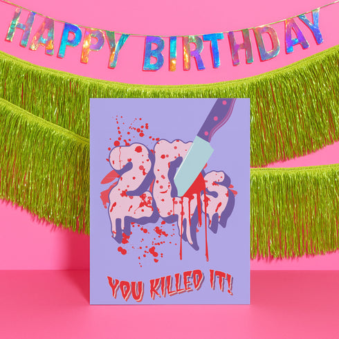 A 5x7 birthday card with a horror-comedy design. The front features the word "20s" in pink gooey lettering with a bloody knife stabbing through it, surrounded by red blood splatter. Below it reads "YOU KILLED IT!" in dripping red font, set against a purple background with a rainbow foil tinsel curtain behind it. Inside, the right panel says "HERE'S TO THE NEXT VICTIM: YOUR THIRTIES!" with dripping red blood at the top and a pink cherry garland background.