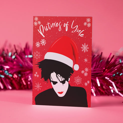 A festive greeting card featuring an illustration of Robert Smith from The Cure, wearing a Santa hat. The card has a red background with white snowflakes and the text "Pictures of Yule" at the top, playfully referencing the song "Pictures of You." The card merges gothic and holiday themes for a unique seasonal twist.