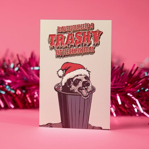 A playful holiday card featuring a raccoon wearing a Santa hat, popping out of a trash can with a mischievous grin. The text above reads "Have Yourself a Trashy Little Christmas" in bold, dripping letters. The card combines humor with a grungy, festive theme for a lighthearted take on the holiday season.