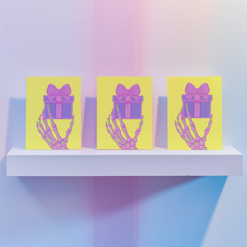 Birthday card with vibrant yellow background. A pink and green skeletal hand holds a purple and pink gift box with large pink bow. The illustration blends festive and spooky elements, creating a playful Halloween-meets-birthday aesthetic. The stylized, colorful design uses contrasting bright colors for a eye-catching effect. Inside message reads: 'Unwrap the Magic. Embrace the Spook. It's Your Day to Shine!' - combining celebratory spirit with a touch of the macabre for a unique birthday greeting.