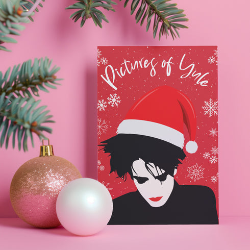 A festive greeting card featuring an illustration of Robert Smith from The Cure, wearing a Santa hat. The card has a red background with white snowflakes and the text "Pictures of Yule" at the top, playfully referencing the song "Pictures of You." The card merges gothic and holiday themes for a unique seasonal twist.