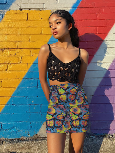 High-waisted bodycon mini skirt with an allover print of rainbow monarch butterflies and colorful tropical flowers on a dark background. Soft, stretchy poly-spandex fabric hugs the body for a flattering fit. Styled for rave, Y2K, and festival outfits. Sizes XS to XL.