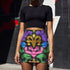 A high-waisted bodycon mini skirt for women shown in two fabric options. The skirt features a vibrant, colorful, symmetrical abstract print with psychedelic, groovy, baroque-inspired shapes in purple, pink, gold, green, and blue on a black background. One version is a smooth, stretchy signature performance knit that is UV reactive and glows under black light. The second version is a glitter knit with a metallic, shimmery foil finish. The mid-thigh length skirt has a tight, curve-hugging fit.