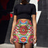 A vibrant bodycon mini skirt featuring a structured grid of retro geometric medallions. The pattern includes concentric circles in red, yellow, and blue, stylized daisy flower motifs, and red and white checkered squares. The skirt has a high-waist elastic band and a smooth, matte performance knit texture. The background is a neutral off-white. This maximalist design utilizes saturated primary colors to create a 70s-inspired psychedelic aesthetic. No purple or navy is present in the print.