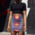 Form-fitting bodycon mini skirt with a mid-rise elastic waistband, falling to mid-thigh in smooth stretch fabric. It features a bold groovy mod floral print: oversized orange flowers with white inner petals and golden yellow centers, black flower accents, and white four-point sparkles, all set on a deep purple background. Rainbow borders in blue, green, orange, and yellow run down each side. The all-over design is a flat dye-sublimated print, available in a smooth knit or glitter knit finish.