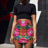 Form-fitting bodycon mini skirt with a mid-rise elastic waistband, falling to mid-thigh in smooth stretch fabric. It features a bold 70s pop art floral print: oversized hot pink flowers with cream scalloped centers, royal blue middles, and fine purple rays, beside a flaming orange tulip, royal blue geometric shapes, and olive green leaves on a black background. The all-over design is a flat dye-sublimated print in vibrant, high-contrast color, available in a smooth knit or glitter knit finish.
