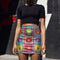 Form-fitting bodycon mini skirt with a mid-rise elastic waistband, falling to mid-thigh in smooth stretch fabric. It features a bold retro patchwork floral print: large mod daisies in red, purple, yellow, and green, set on a sky blue background with scattered green leaves. A central column frames single flowers on purple and pink tiles, edged by red and pink beaded borders. The all-over design is a flat dye-sublimated print, available in a smooth knit or glitter knit finish.
