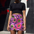 A high-waisted, bodycon mini skirt with a vibrant psychedelic floral pattern. The curve-hugging skirt features a mid-thigh length and black stitching at the waistband and hem. The all-over print consists of abstract, fuzzy-edged floral bursts in electric colors, including hot pink, magenta, neon green, and electric purple, set against a black background. The skirt is shown in two finishes: a smooth, matte signature knit and a metallic glitter knit with a reflective silver foil.