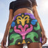 A high-waisted bodycon mini skirt for women shown in two fabric options. The skirt features a vibrant, colorful, symmetrical abstract print with psychedelic, groovy, baroque-inspired shapes in purple, pink, gold, green, and blue on a black background. One version is a smooth, stretchy signature performance knit that is UV reactive and glows under black light. The second version is a glitter knit with a metallic, shimmery foil finish. The mid-thigh length skirt has a tight, curve-hugging fit.