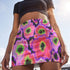 A high-waisted, bodycon mini skirt with a vibrant psychedelic floral pattern. The curve-hugging skirt features a mid-thigh length and black stitching at the waistband and hem. The all-over print consists of abstract, fuzzy-edged floral bursts in electric colors, including hot pink, magenta, neon green, and electric purple, set against a black background. The skirt is shown in two finishes: a smooth, matte signature knit and a metallic glitter knit with a reflective silver foil.