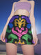 A high-waisted bodycon mini skirt for women shown in two fabric options. The skirt features a vibrant, colorful, symmetrical abstract print with psychedelic, groovy, baroque-inspired shapes in purple, pink, gold, green, and blue on a black background. One version is a smooth, stretchy signature performance knit that is UV reactive and glows under black light. The second version is a glitter knit with a metallic, shimmery foil finish. The mid-thigh length skirt has a tight, curve-hugging fit.