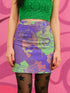 High-waisted bodycon mini skirt with a bold floral collage print in green, purple, and pink. Features comic-style roses and dot patterns on stretchy polyester-spandex fabric. Styled for festivals and summer parties. Available in sizes XS to XL.