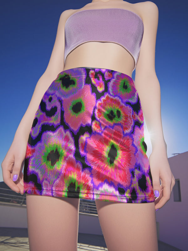 A high-waisted, bodycon mini skirt with a vibrant psychedelic floral pattern. The curve-hugging skirt features a mid-thigh length and black stitching at the waistband and hem. The all-over print consists of abstract, fuzzy-edged floral bursts in electric colors, including hot pink, magenta, neon green, and electric purple, set against a black background. The skirt is shown in two finishes: a smooth, matte signature knit and a metallic glitter knit with a reflective silver foil.