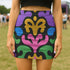 A high-waisted bodycon mini skirt for women shown in two fabric options. The skirt features a vibrant, colorful, symmetrical abstract print with psychedelic, groovy, baroque-inspired shapes in purple, pink, gold, green, and blue on a black background. One version is a smooth, stretchy signature performance knit that is UV reactive and glows under black light. The second version is a glitter knit with a metallic, shimmery foil finish. The mid-thigh length skirt has a tight, curve-hugging fit.